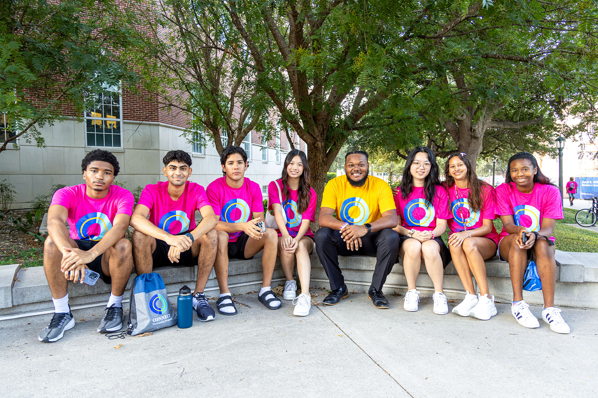 group of people in pink and yellow t shirts sitting on bench outside in front of trees