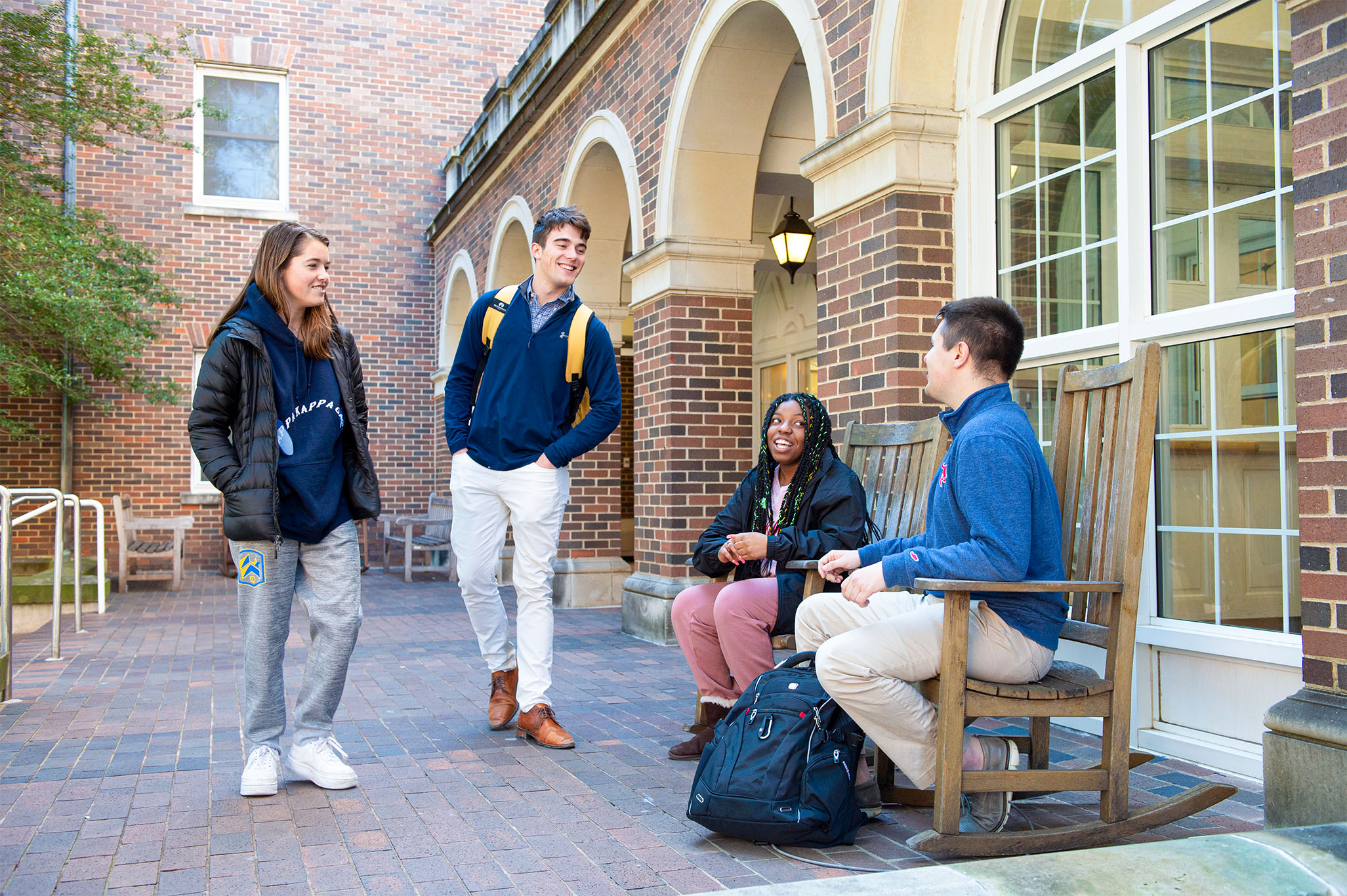 Four students smiling and talking outside of brick building, two sitting in chairs and two standing