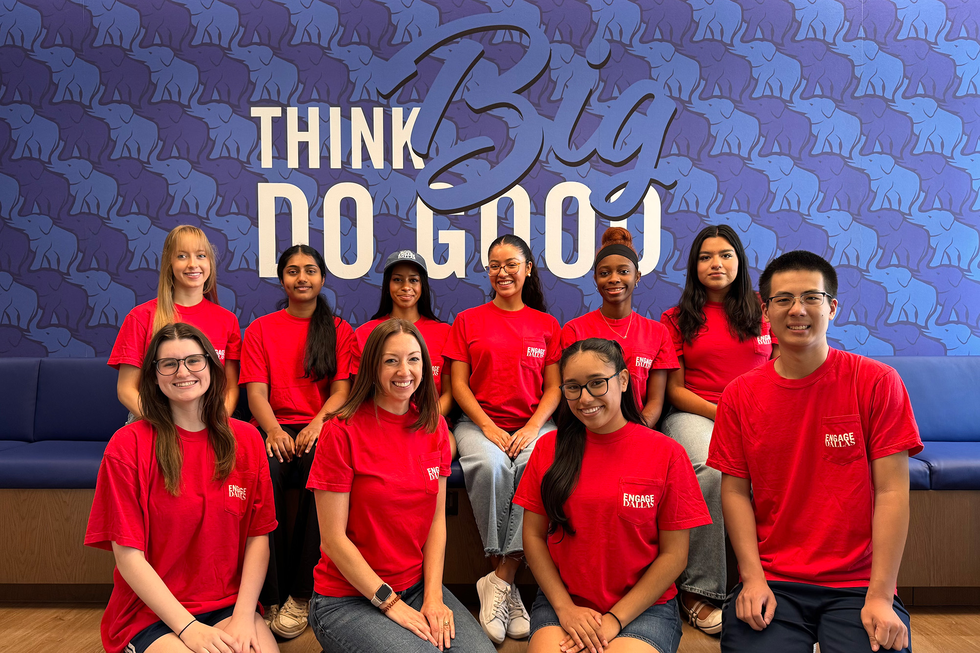 group of people posing for photo in red shirts sitting on bench in front of dark and light blue designed wall