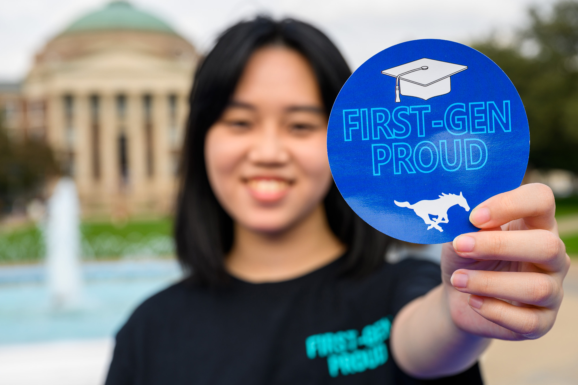 girl in black shirt holding first-gen proud sticker in focus outside with building in background