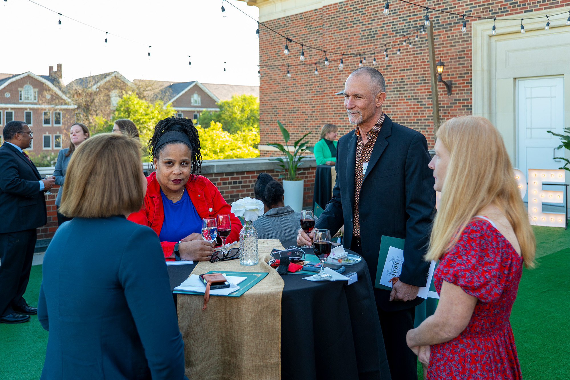 4 people standing outside at table talking with brick wall and string lights in the background