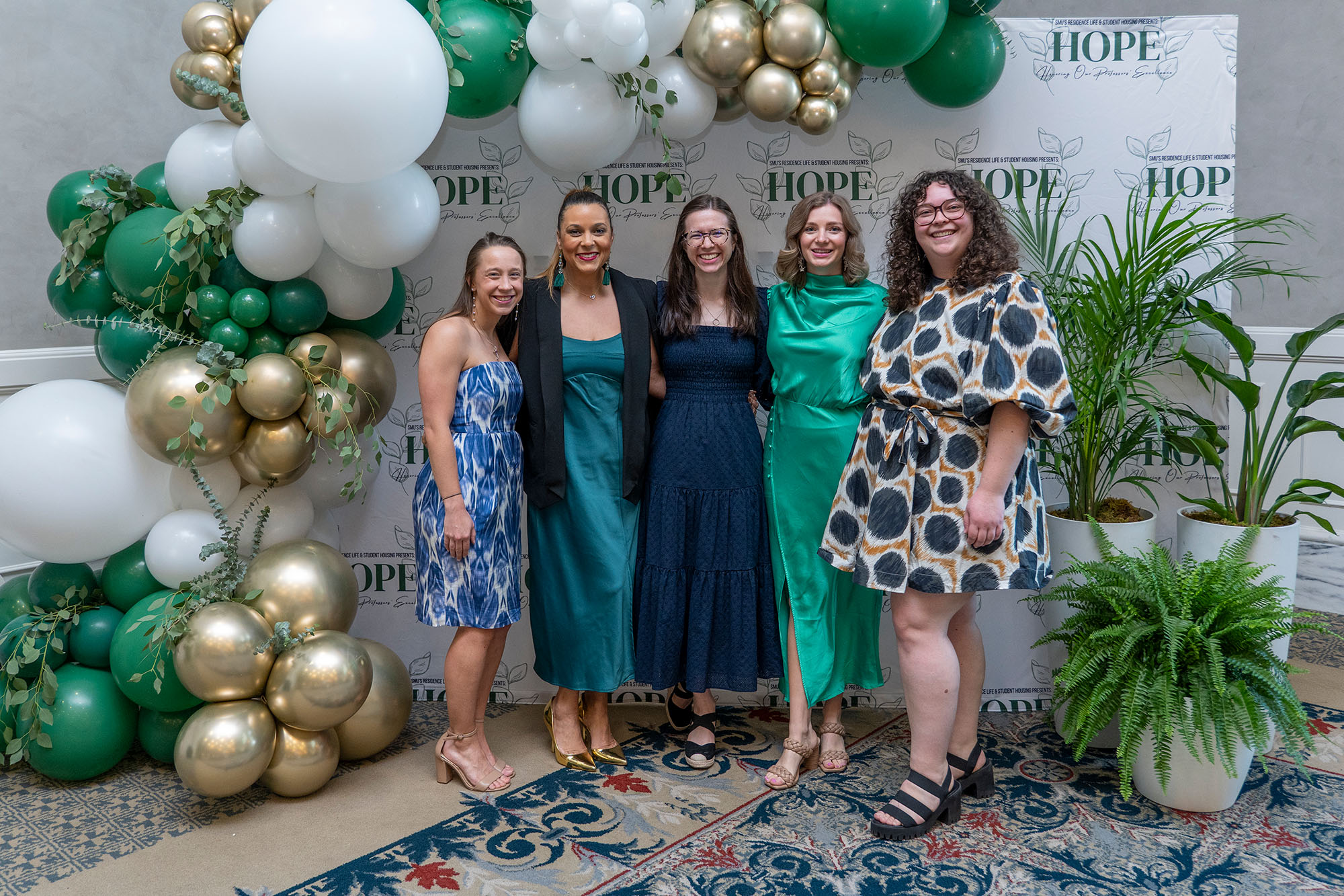 five people posing for photo in front of white backdrop with HOPE written out in green letters and repeating, white, gold, and green balloons on the left, three plants of varying sizes on the right