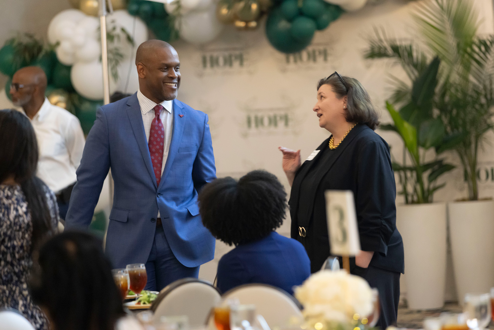 two people standing and talking by tables with green and white balloons in background