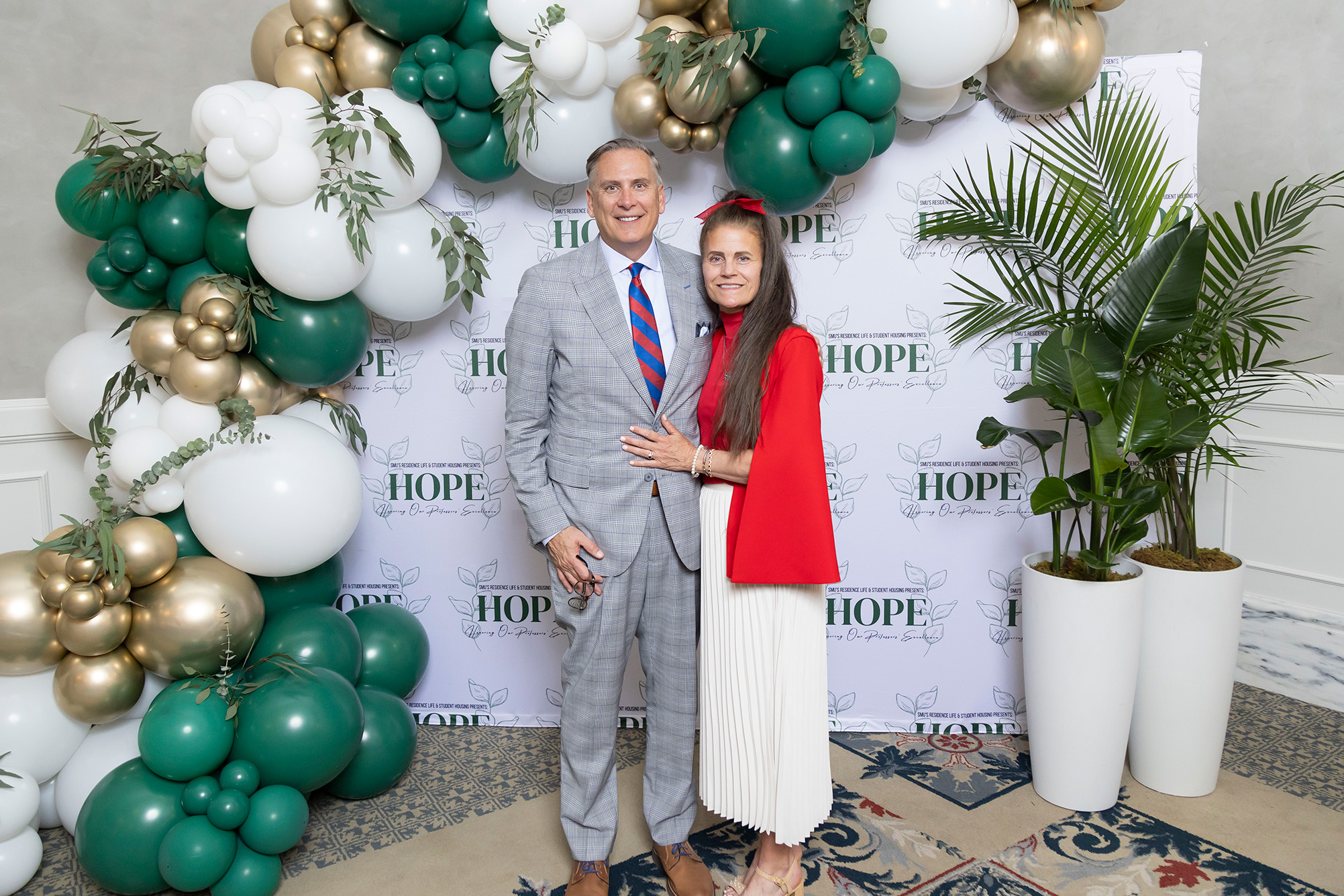 two people posing for photo with green, white, and gold balloons and trees in background