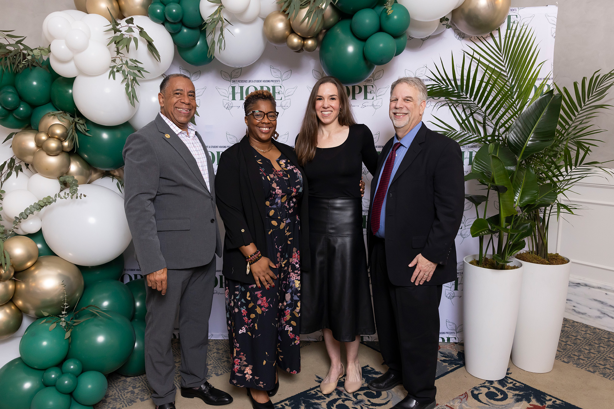 four people posing for photo with green, white, and gold balloons and trees in background