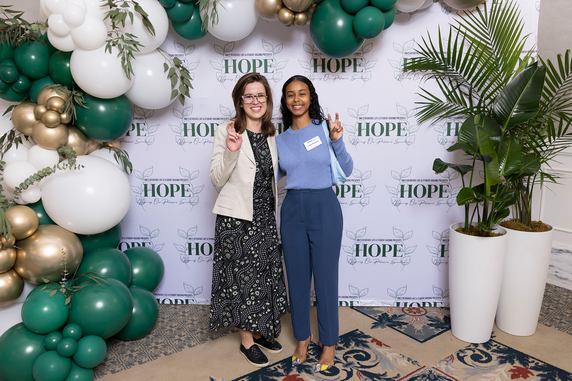 two people posing for photo with green, white, and gold balloons and trees in background