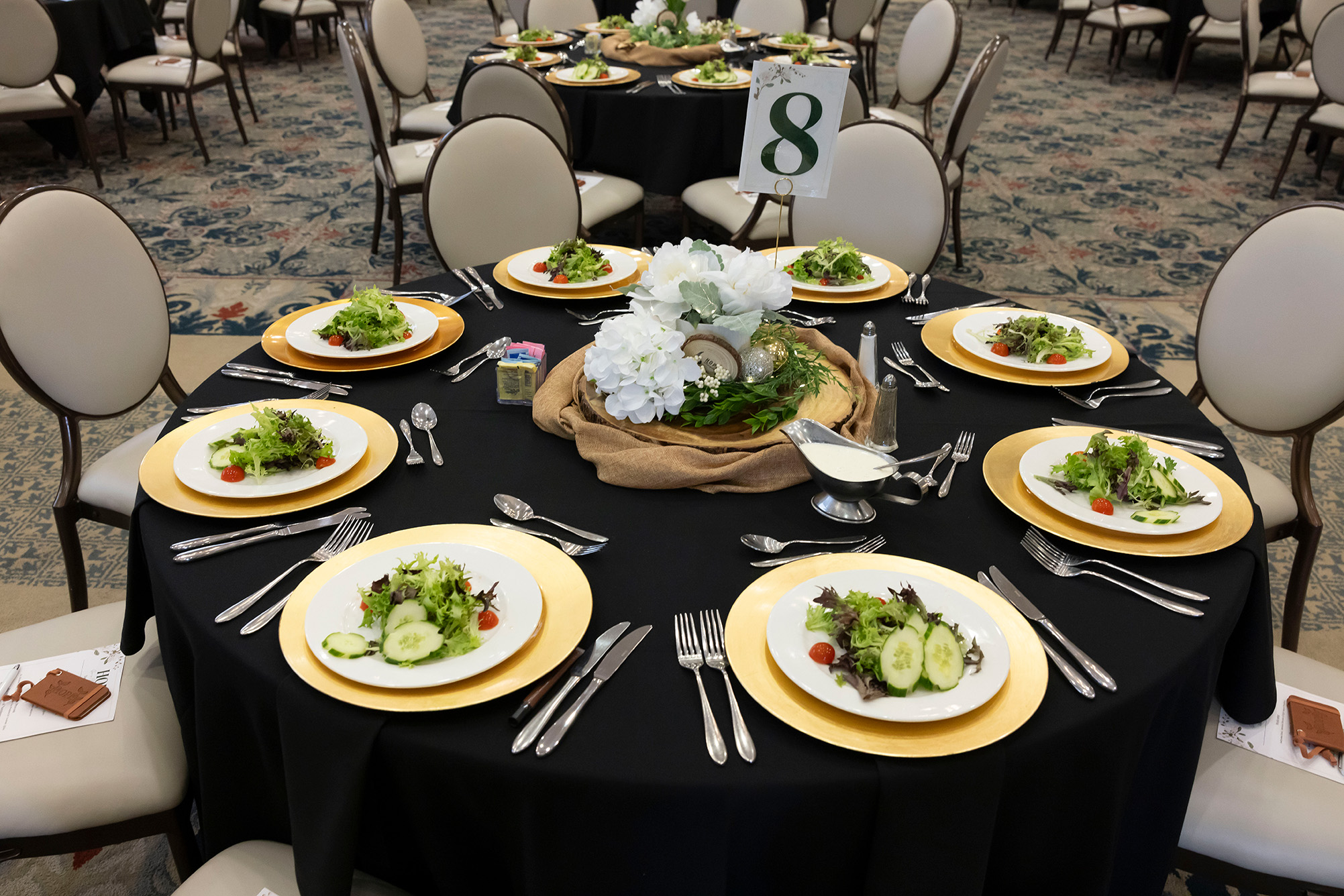 table set with black tablecloth, white and gold dishes, white and green floral center piece, with white and black chairs