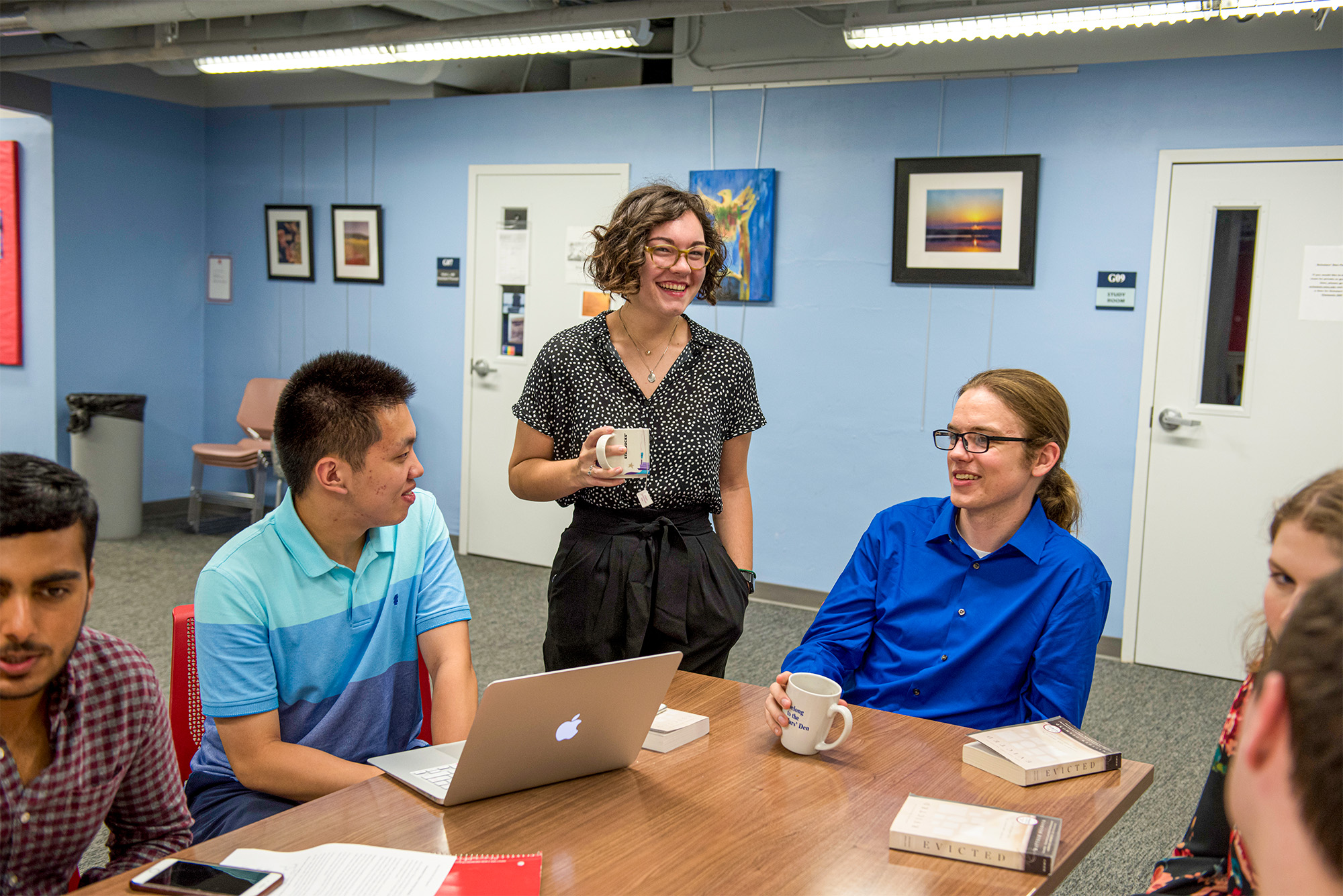 students sitting at table and one person standing with coffee cup in hand, talking and smiling