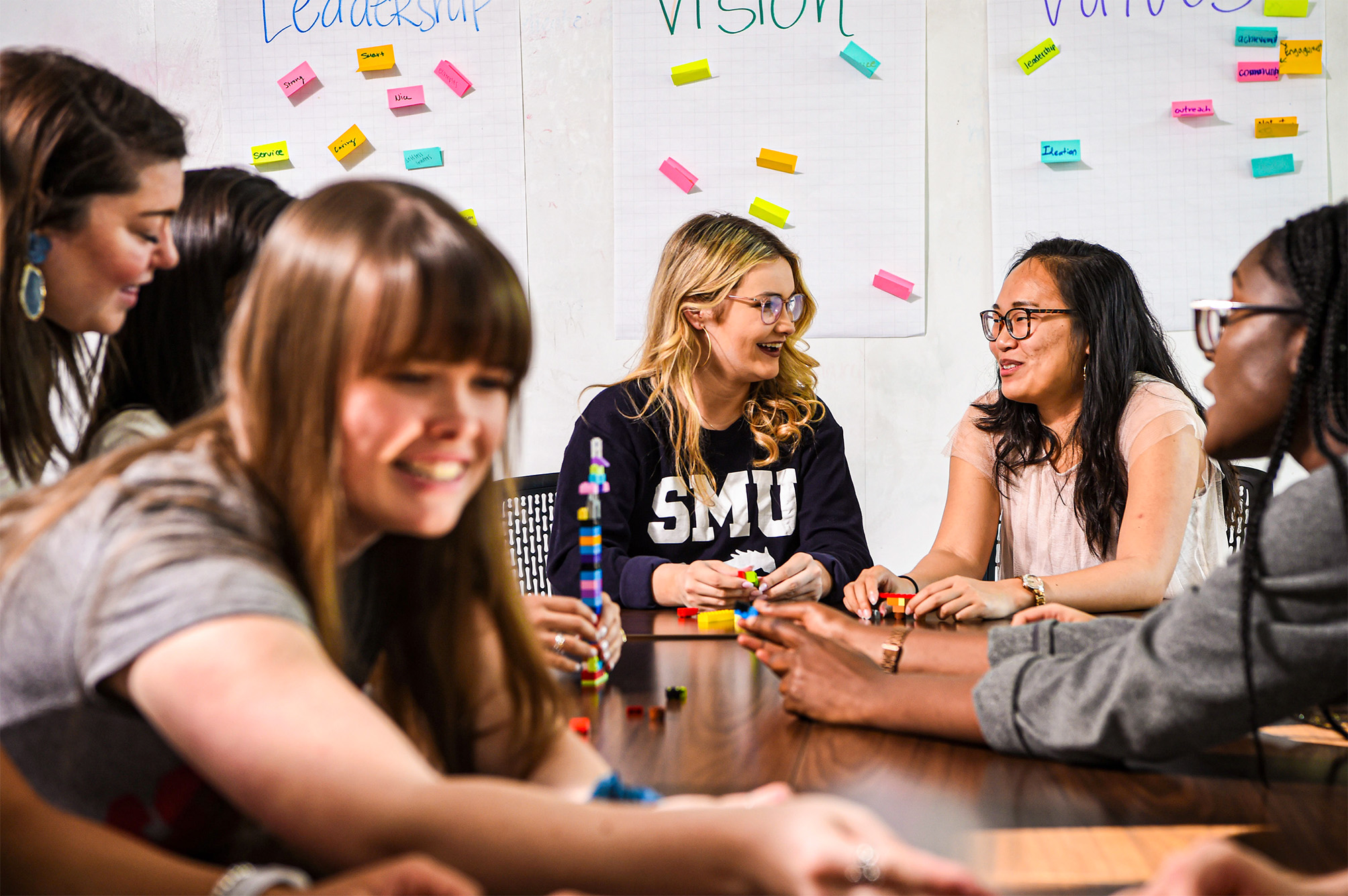 students sitting at table talking with whiteboard in background with different color post-it notes on the board