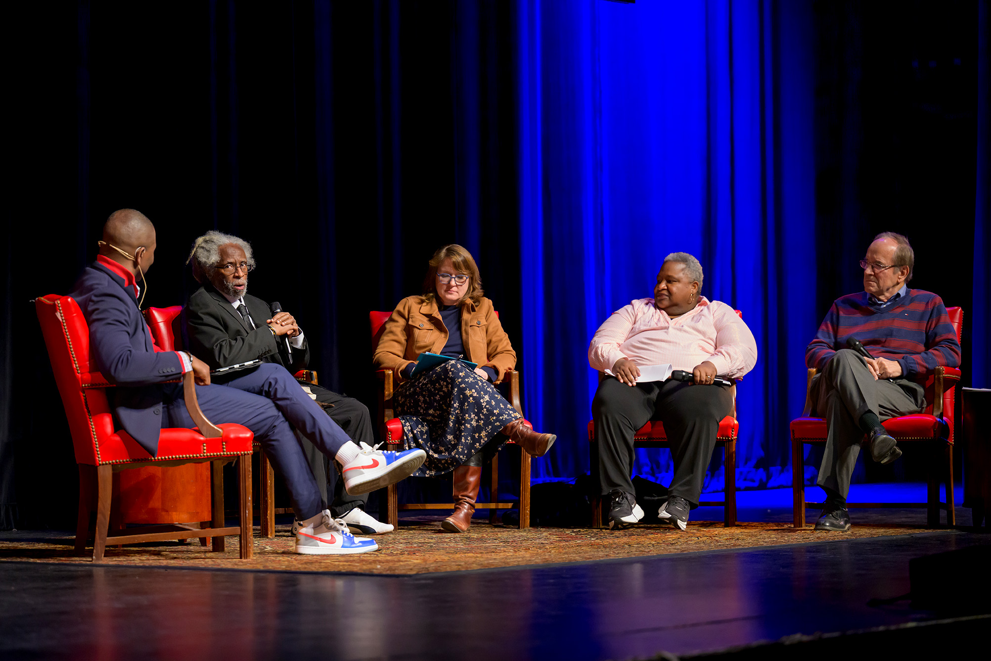 five people sitting in chairs on stage with microphones for a panel discussion with a black stage floor, blue curtains in background