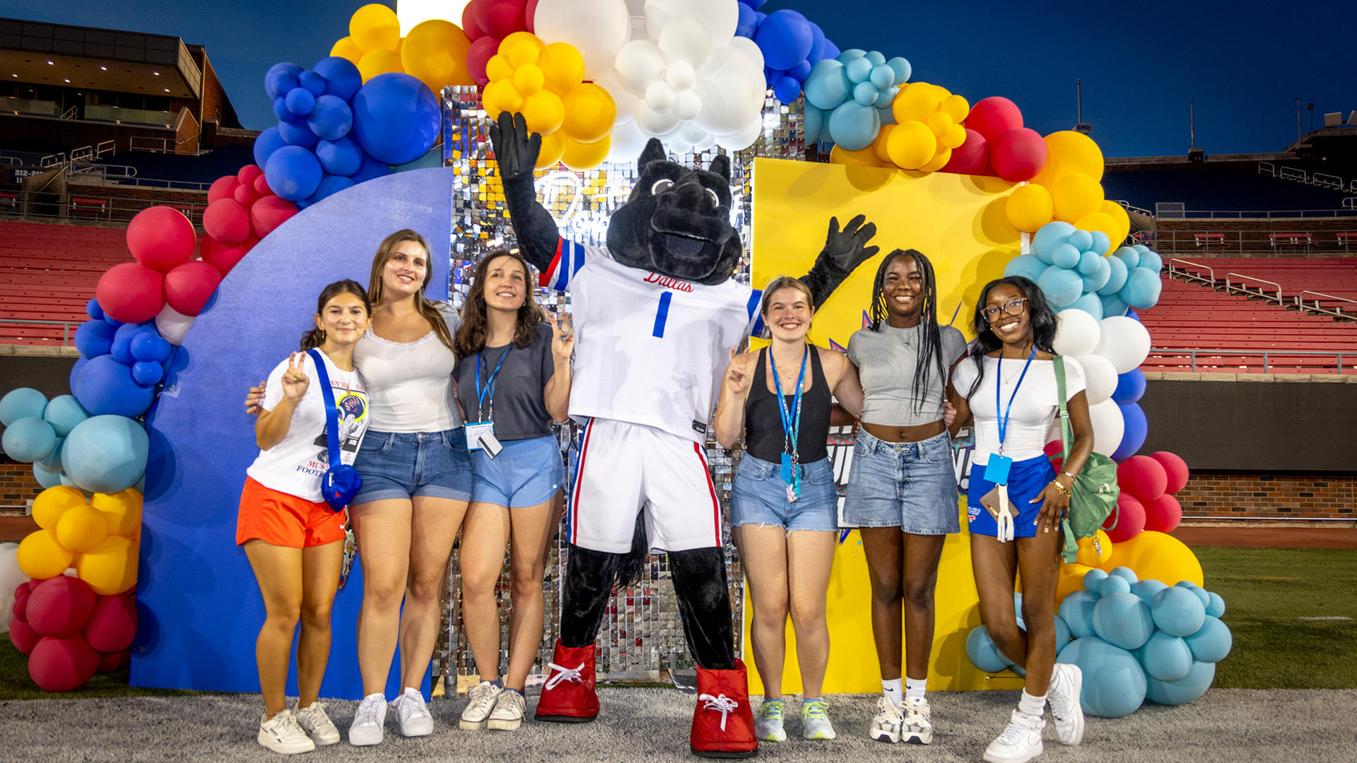 six students standing with peruna mascot outside in front of blue and yellow wooden arched backdrop with red, blue, white, yellow, and teal balloons