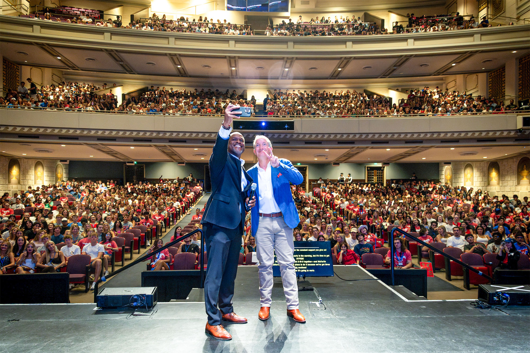 two people on a stage taking a selfie with large group of people inside