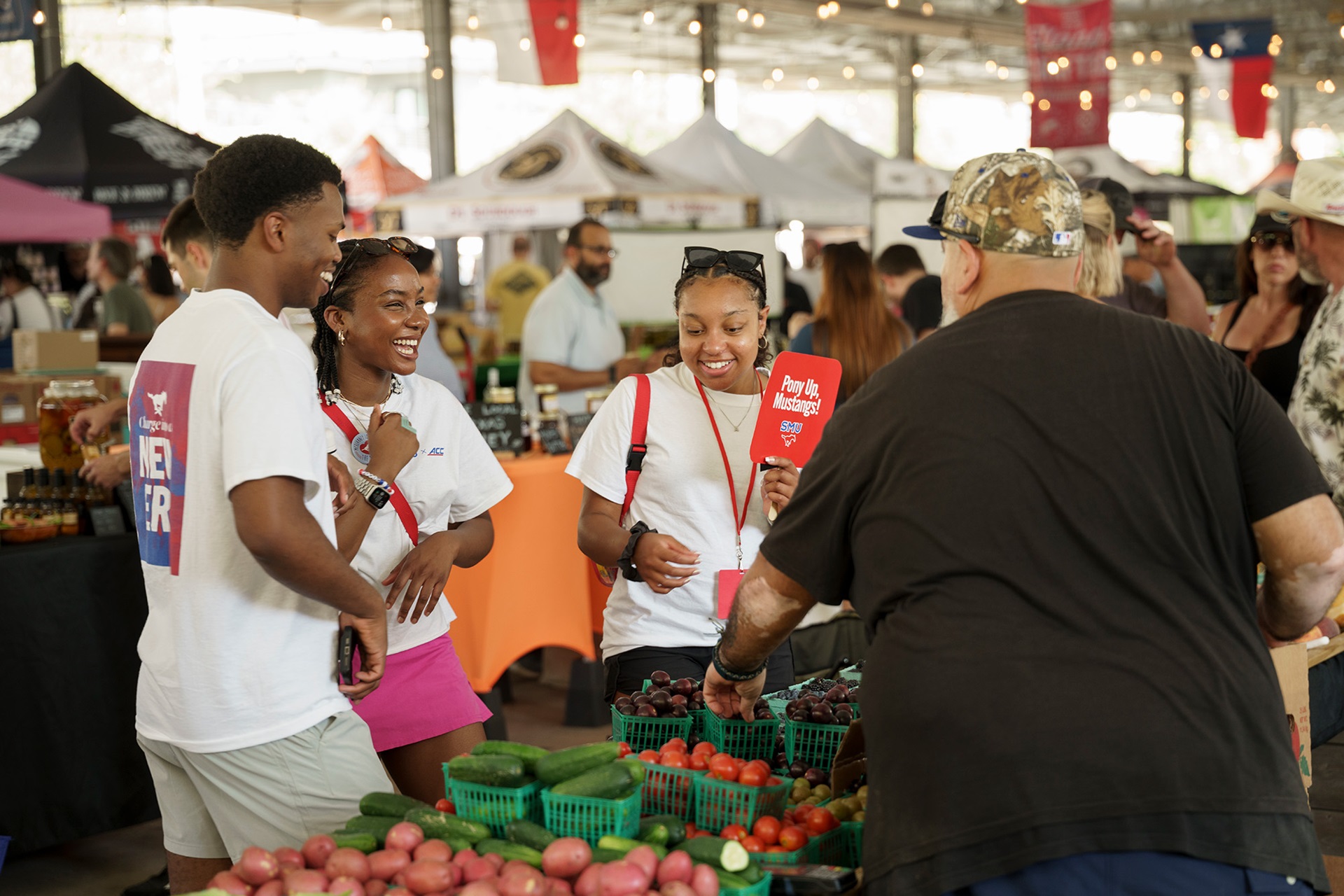 three students in white shirts talking to vendor in a black shirt at looking at vegetables at the farmers market