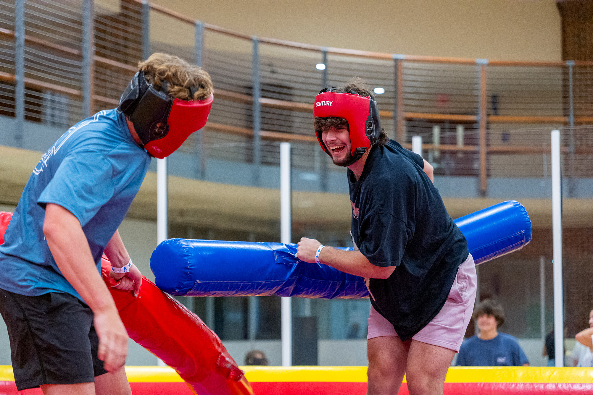 two students playing with large foam sticks and read foam helmets indoors