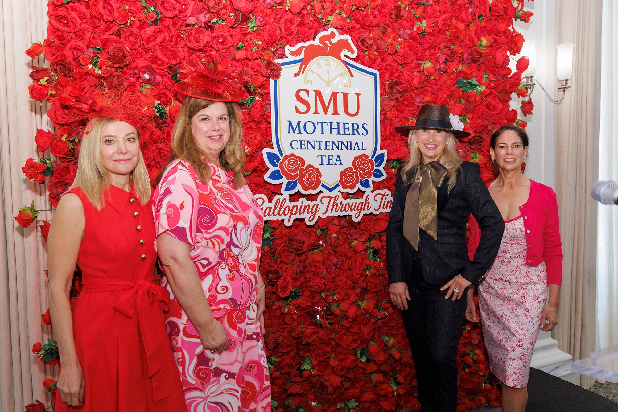women in pink, red, and black dresses standing in front of wall of red roses posing for photo