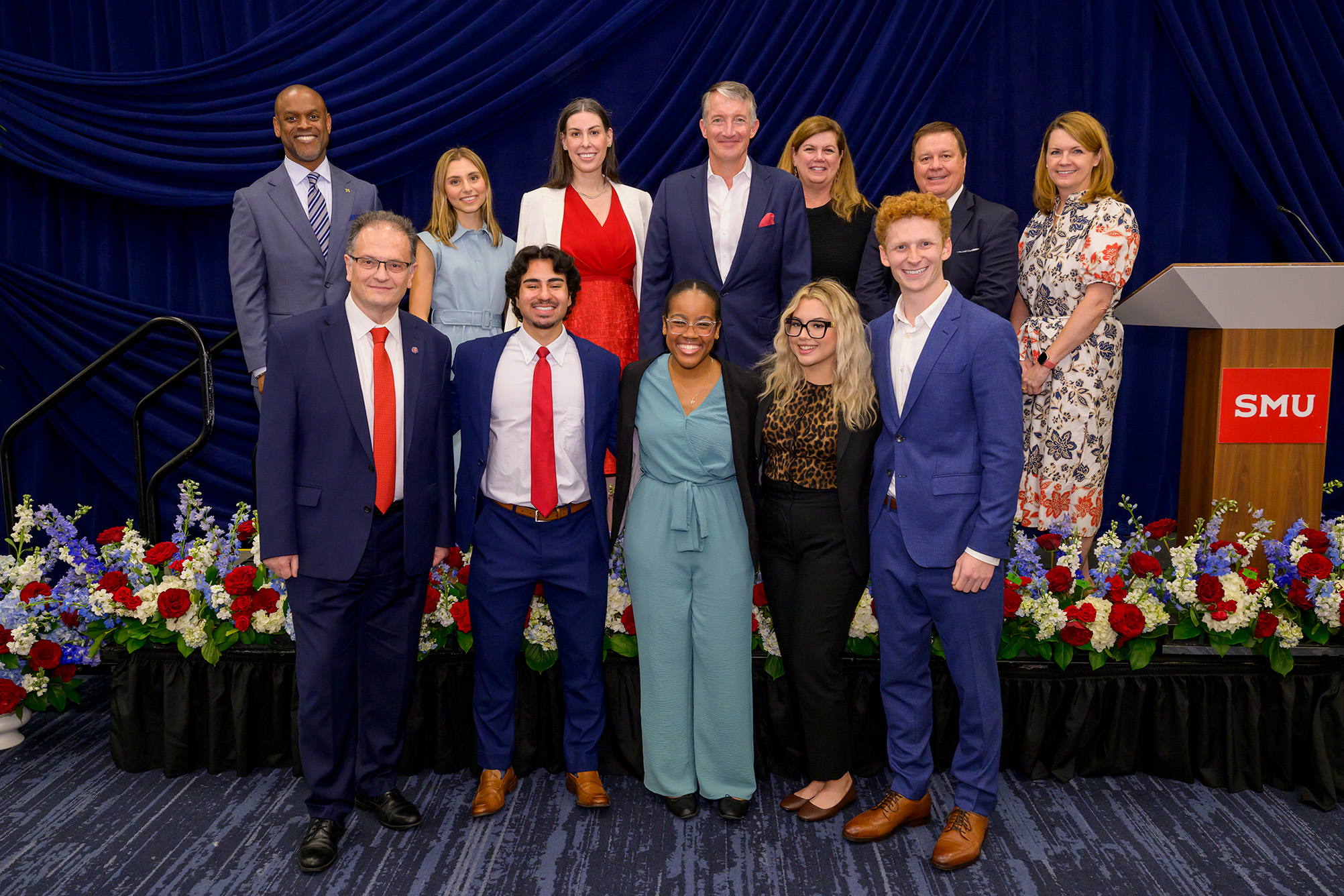 group of people indoors standing in front of dark blue curtains posing for photo with red, blue, and white flower arrangements