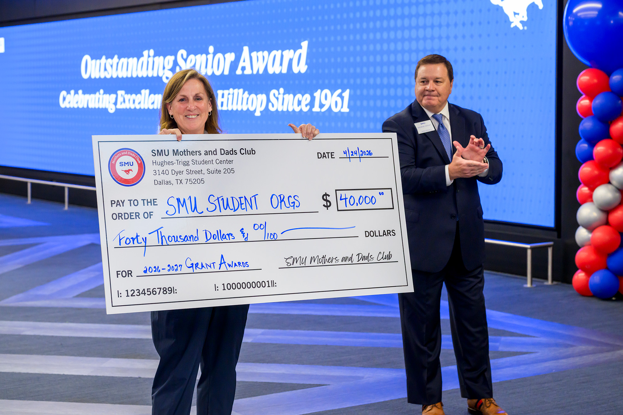 two people standing inside, person on left holding large check indicating a donation to student organizations