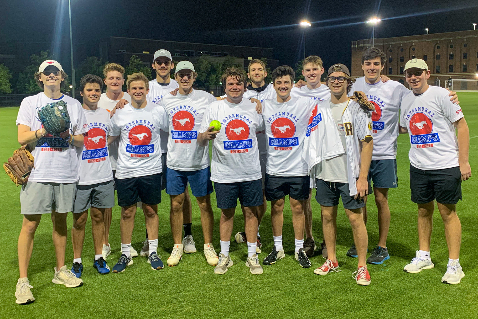 group of college men in white and red tshirts standing outside at night on turf field with softball gloves posing for photo