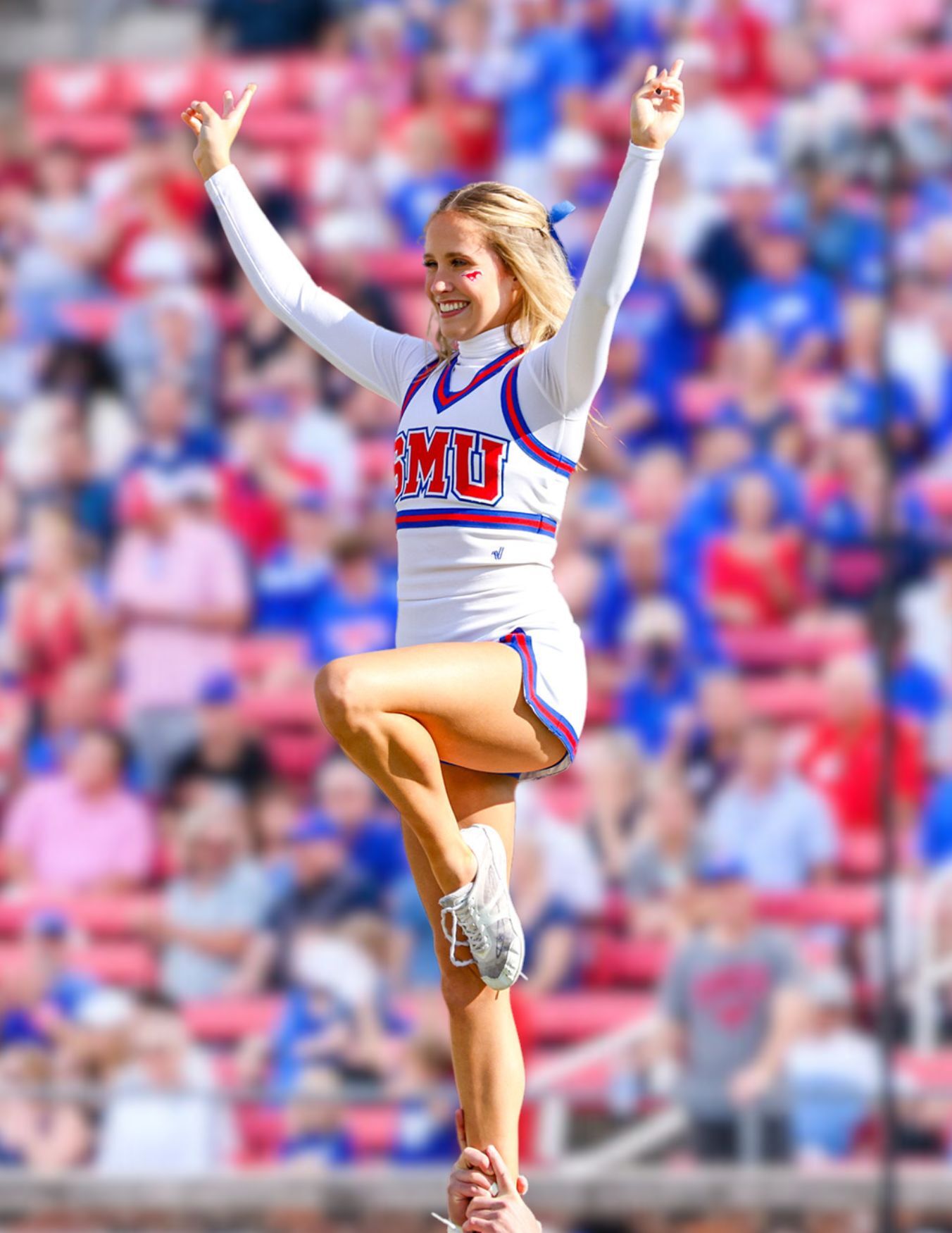 SMU cheerleader lifted in the air on one leg in white long sleeve uniform, red SMU logo and blue trim on uniform, holding pony up hand signal, background is people sitting in stands wearing red, blue, and white