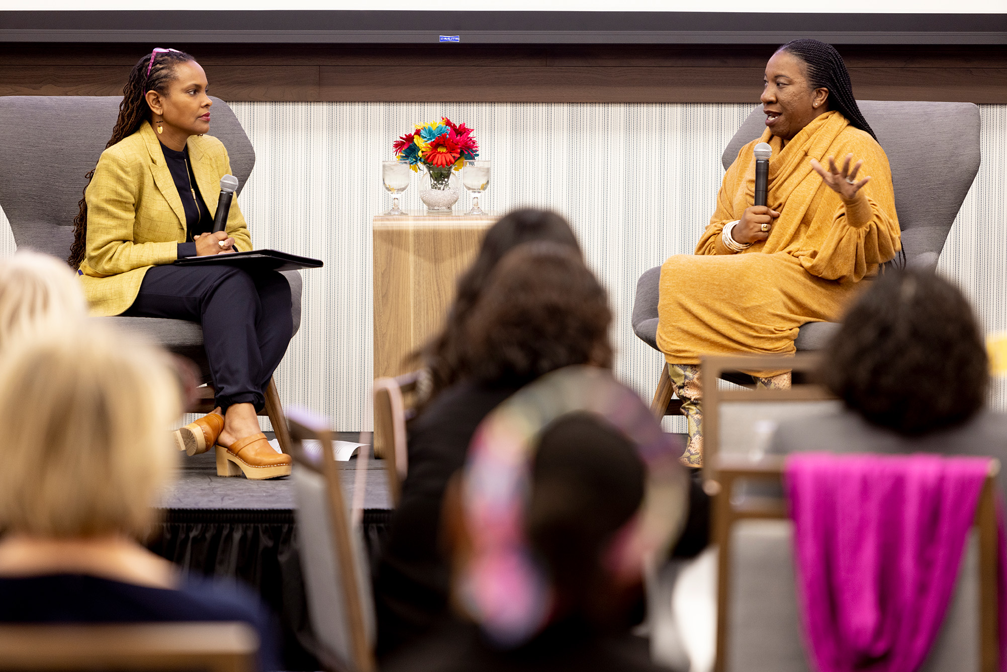 two people sitting in grey chairs on stage talking with microphones