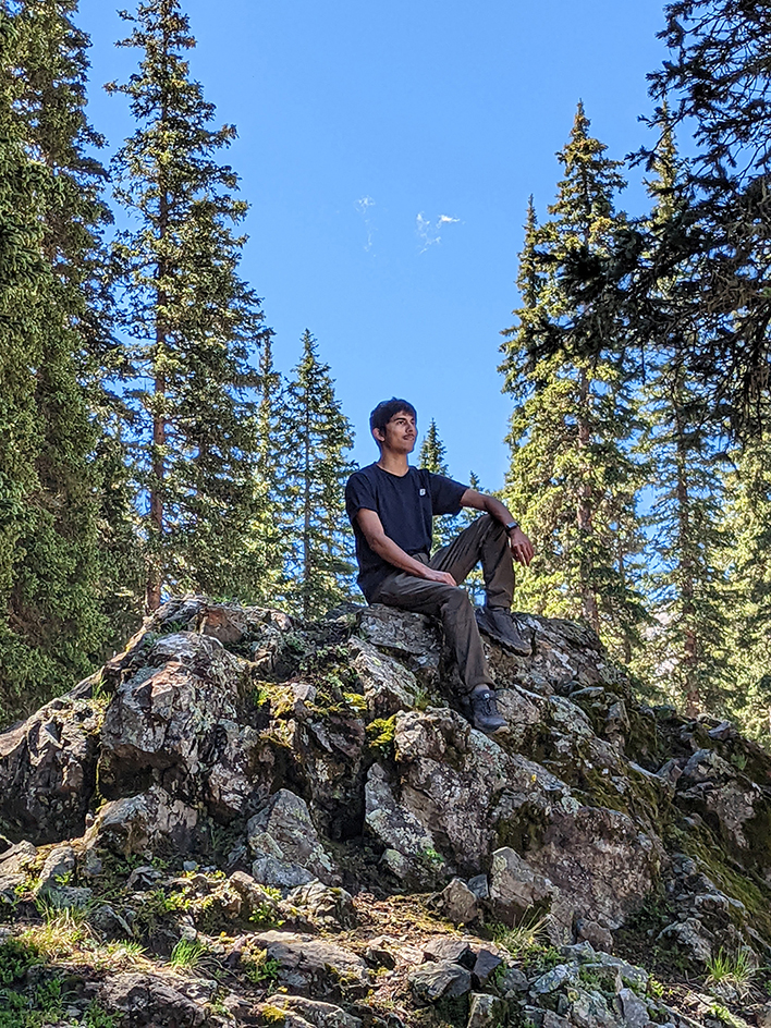 Taos student Aria Shame sits atop a rock surrounded by tall trees in Taos, New Mexico.