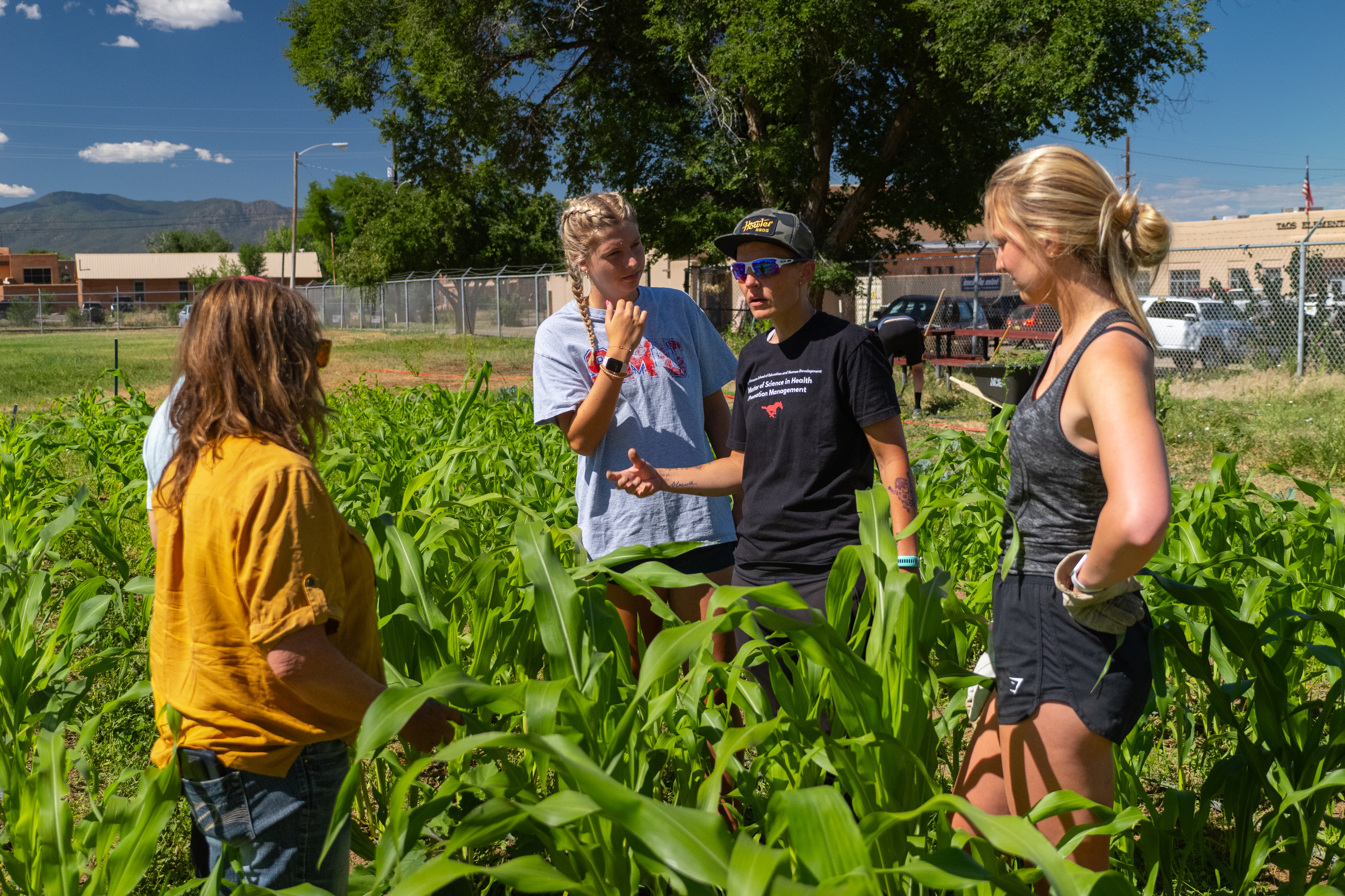 Group of people standing in a garden of waist high crops - all are looking at one person in center of group who is gesturing while speaking. 