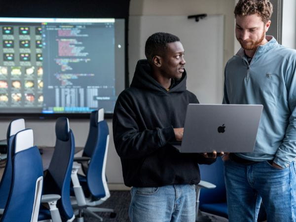 Two students looking at a laptop in a classroom in front of a projection screen with data on it