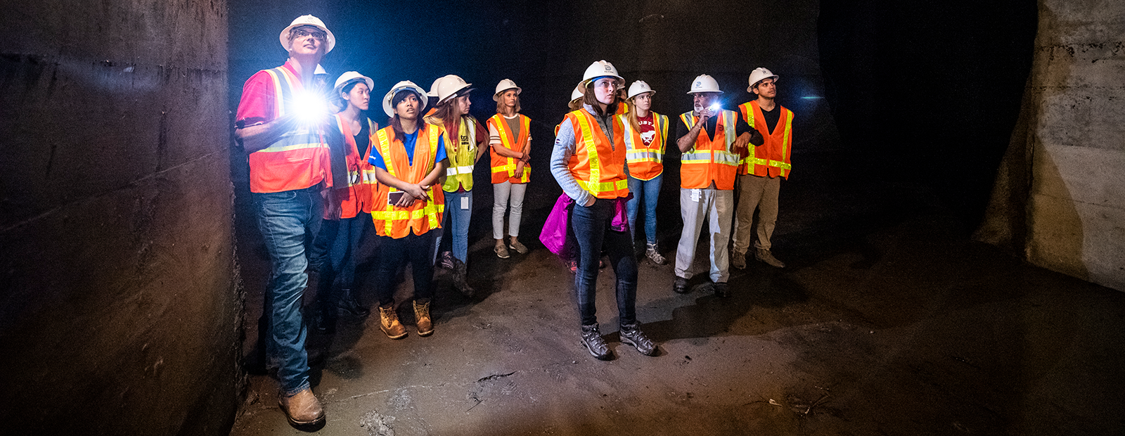 A group of civil engineering students with hard hats and safety vests are exploring a tunnel with flashlights.